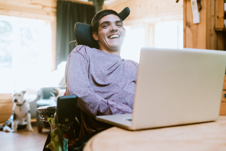 Ein behinderter junger Mann sitzt in seinem Rollstuhl an einem Schreibtisch mit einem aufgeklapptem Laptop und blickt lachend in die Kamera.
