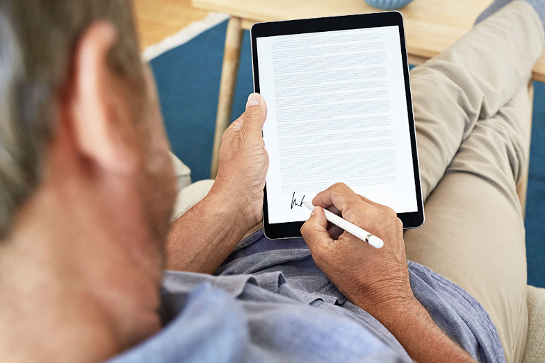 Symbolfoto digitale Signatur: Mann sitzt auf dem Sofa und unterschreibt digital auf einem Tablet