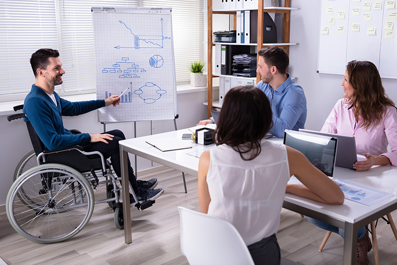 Symbolbild Diversität: Mann im Rollstuhl hält eine Präsentation und zeigt etwas auf einem Flipchart. Seine Kolleginnen und Kollegen hören ihm aufmerksam zu.