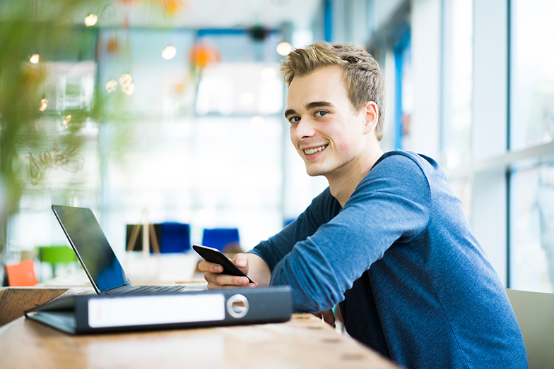Symbolbild: Junger Student sitzt am Tisch mit Studienmaterial, Laptop und hält ein Smartphone in der Hand (Quelle: Getty Images / Westend61) Symbolbild: Junger Student sitzt am Tisch mit Studienmaterial, Laptop und hält ein Smartphone in der Hand