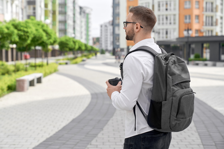 Symbolbild: Junger bärtiger Mann mit Rucksack und Thermobecher in Seitenansicht mit Blick auf mehrstöckige Gebäude. (Quelle: 123rf / serhiibobyk) Symbolbild: Junger bärtiger Mann mit Rucksack und Thermobecher in Seitenansicht mit Blick auf mehrstöckige Gebäude.