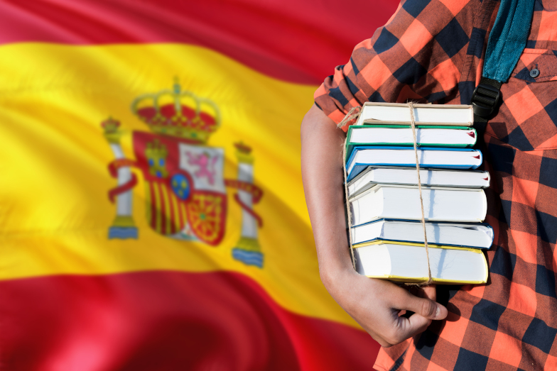 Person hält vor großer spanischer Nationalflagge Bücherstapel unter dem Arm. (Quelle: Adobe Stock / sezerozger) Person hält vor großer spanischer Nationalflagge Bücherstapel unter dem Arm.